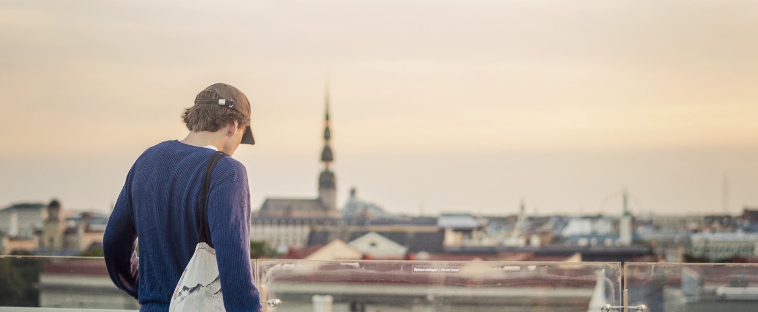 Молодой человек стоит на крыше здания CENTRUS и любуется панорамой центра Риги с башнями и крышами.